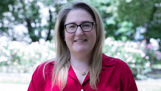 Abby Edwards, Frau mit blonden Haaren, Brille und roter Bluse steht im Park