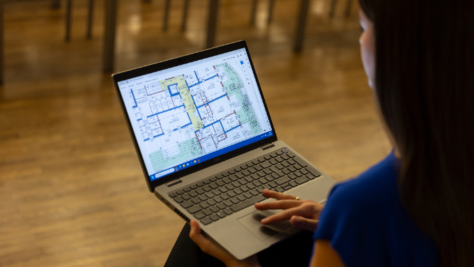 Am Bild zu sehen ist eine sitzende Dame, welche einen Laptop auf ihren Oberschenkeln platziert hat und sich einen Bauplan darauf ansieht.