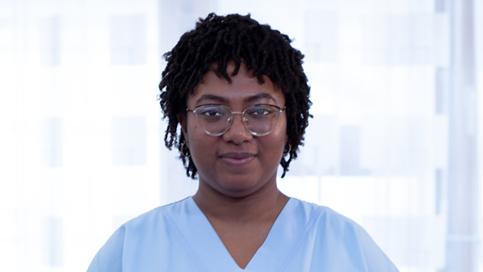 Frau mit dunklen Haaren, Brille und dunklen Augen trägt Pflegegewand und ist im Krankenhaus bei der Arbeit.