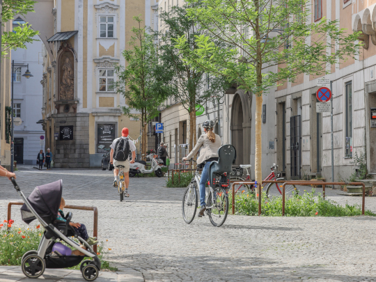 Blick auf den Pfarrplatz auf Seite des Alten Rathauses Richtung Domgasse, zu sehen sind Laubbäume, zwei Personen auf Fahrrädern und ein Kinderwagen.