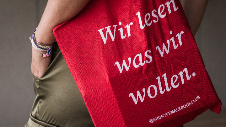 Rote Stofftasche mit weißer Aufschrift 'Wir lesen was wir wollen.' und 'ANGRYFEMALEBOOKCLUB' an der Seite einer Person in olivgrüner Hose mit Hand in Tasche