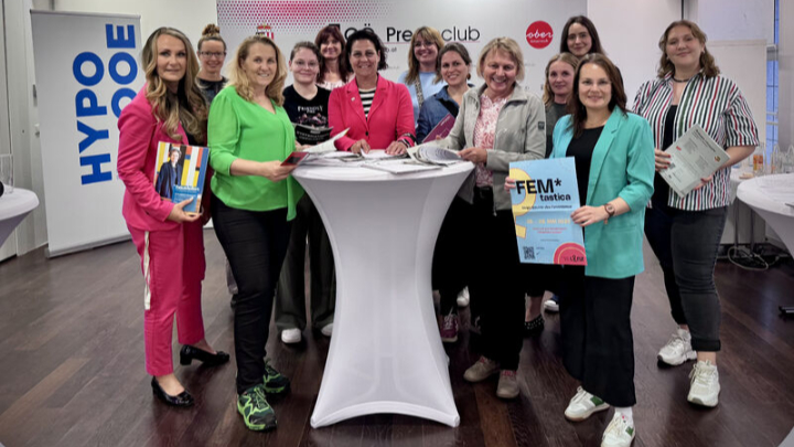 Gruppe von Frauen steht um einen hohen, weißen Stehtisch in einem Raum mit Werbebannern im Hintergrund, einige halten Magazine oder Broschüren