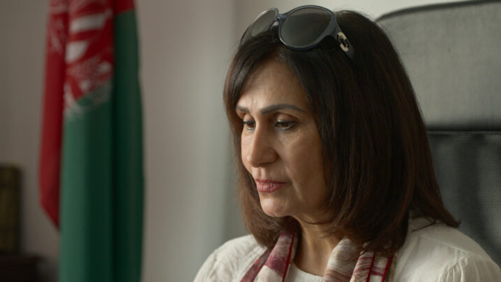 Frau mit mittellangen braunen Haaren, weißem Oberteil und Schal sitzt vor einem afghanischen Flaggenbanner