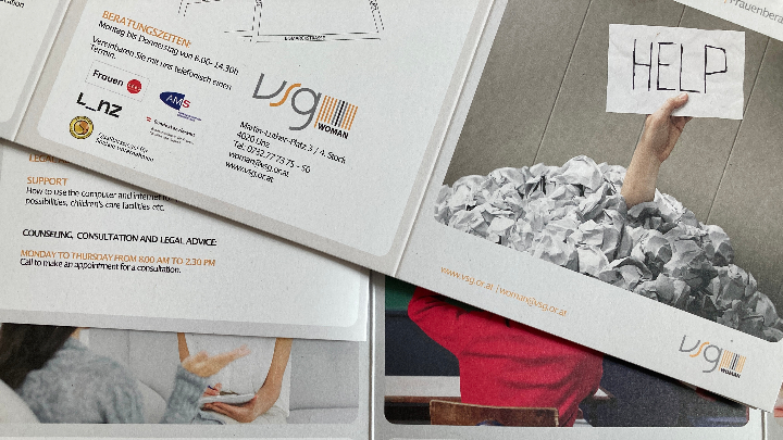 Im Vordergrund ragt ein Arm aus einem Haufen zerknüllter Papierblätter und hält ein Blatt mit der handschriftlichen Aufschrift 'HELP' hoch. Im Hintergrund sind weitere Informationsblätter mit Text und Logos zu sehen, darunter das Logo von VSG Woman