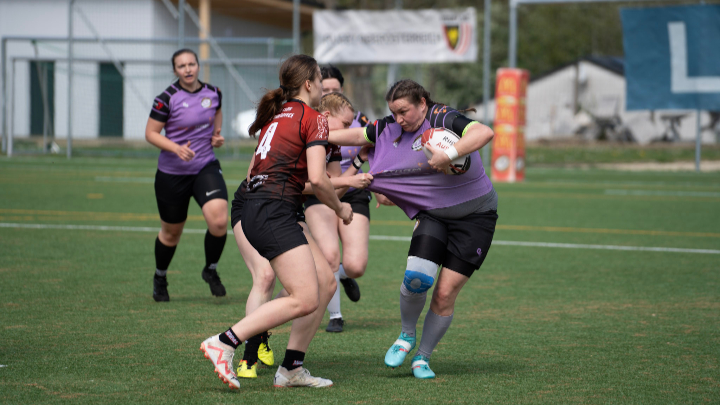 Rugbyspielerin in lila Trikot wird von Spielerin in rotem Trikot am Trikot festgehalten auf Spielfeld