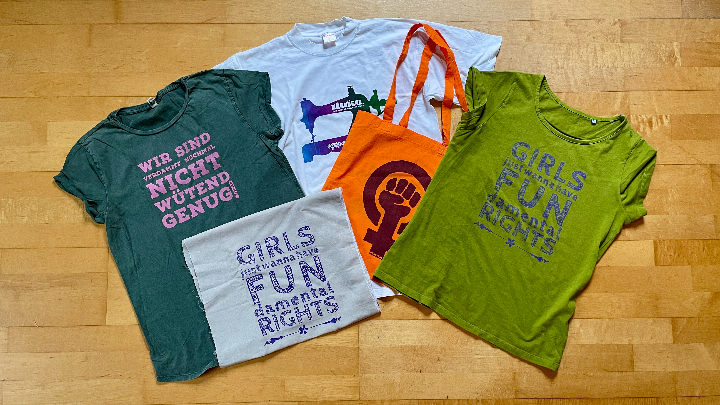 Mehrere T-Shirts und eine Tasche mit feministischen Slogans und Symbolen liegen auf einem Holzboden verteilt