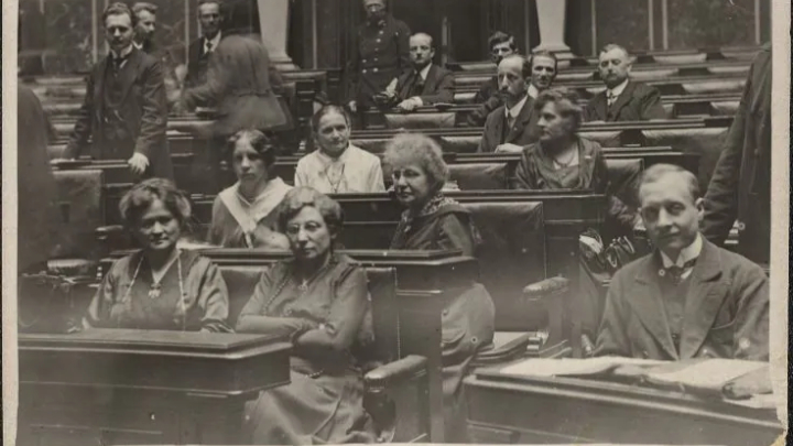 Schwarz-weiß-Fotografie von Parlamentariern und Parlamentarierinnen in einem historischen Sitzungssaal mit Holzpulten und zwei großen Statuen im Hintergrund