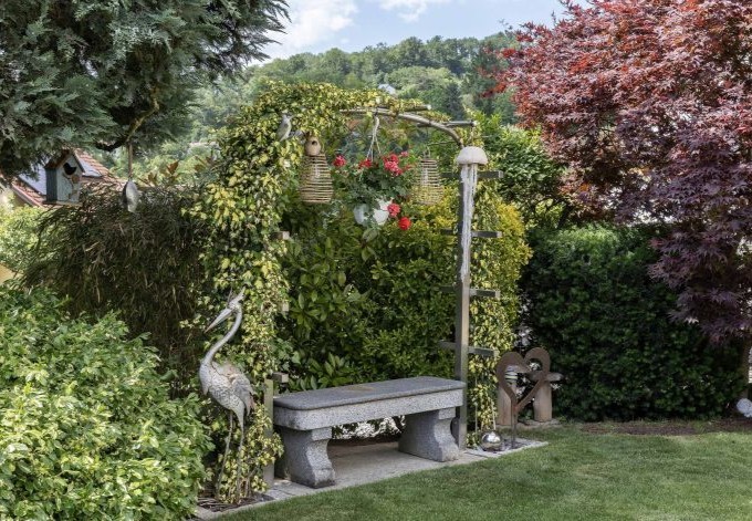 Roman Schinagl Steinbank mit überwachsener Pergola, verschiedene Gehölze und Deko-Reiher